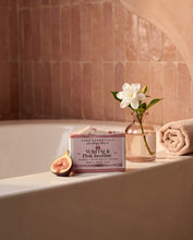 A boxed bath product labeled 'Wild Fig & Pink Jasmine' sits on a bathtub ledge in a warm bathroom setting, with a small glass vase holding a white flower, a sliced fig, and a rolled towel nearby.