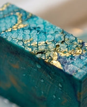 A close-up of a rectangular turquoise and teal textured item with shimmering gold leaf accents and water droplets on its surface.