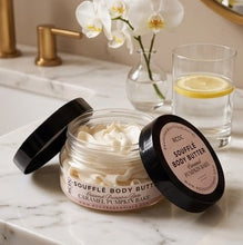 A jar of B2CC Soufflé Body Butter in Caramel Pumpkin Bake, shown open on a white marble countertop with its lid leaning beside it. A glass of water with a lemon slice and white orchids in a vase are softly blurred in the background.