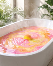 A pink and orange swirling bath bomb explosion in a white bathtub, with a yellow bath bomb floating in the center and steam rising. Green houseplants are visible in the softly lit bathroom background.