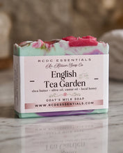 A boxed bar of soap labeled 'English Tea Garden' by RCDC Essentials, shown on a marble surface with a blurred warm-toned background. The soap itself has a pale mint green base with pink and lavender swirls and bright pink top pieces.