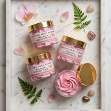 Four jars of whipped soap scrub by Woodstock Cosmetics arranged on a white marble tray with decorative green leaves, pink petals, and stones. One jar is open, showing pink whipped product, while the others are closed with gold lids.