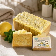 Artisan lemon poppy soap bars displayed on a light stone surface, with a whole lemon, mint leaves, folded towel, and soap packaging visible. The front bar is sliced to show the creamy yellow soap and dark poppy seed inclusions, next to a branded soap box labeled 'Lemon Poppy' by No.