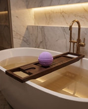 A dark brown wooden bath caddy draped over a white freestanding bathtub, holding a purple bath bomb. The scene is set in a luxurious bathroom with a gold faucet, marble walls, and warm under-cabinet lighting.
