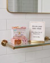 A bar of soap and a honeycomb-shaped honey product from Rose Clay & Honey are displayed on a glass bathroom shelf with a brass holder, next to a sign that reads 'Elevate your daily ritual. Shop Rose Clay & Honey now.