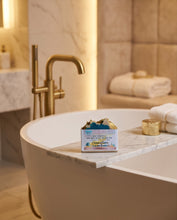 A boxed bath product labeled 'Coconut Love Bath Soak' sits on the edge of a freestanding bathtub in a warm, modern bathroom with gold fixtures and folded white towels.