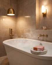 A freestanding white bathtub filled with steaming water in a modern bathroom, with a pink patterned soap bar resting on a small marble tray beside the tub. The room features warm beige tile walls, a gold pendant light, and a wall-mounted sconce.