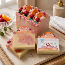 A photo of bars of handmade soap decorated with raspberry and tangerine designs, with a slice of the bar showing the swirled pink and white colors. The soap is accompanied by decorative flowers and the packaging is visible.