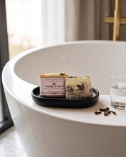 A bar of soap from Wood Essentials 'Orange & Clove' sits on a black oval tray on the rim of a white freestanding bathtub, next to a glass of water and a few cloves.