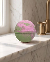 A spherical bath bomb with a pink and green marbled pattern sits on a white marble countertop next to a gold faucet in a bathroom setting.