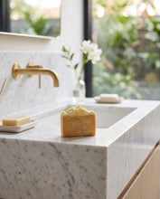 A rectangular bar of yellowish soap with visible speckles sits on a white marble countertop in a modern bathroom setting with a brass faucet and a blurred window view in the background.