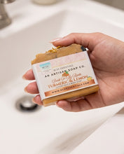 A hand holding a bar of organic artisan soap from RCDC Essentials, featuring a white label with orange and brown text that reads 'Turmeric & Lemon'. The soap bar itself is a warm golden-brown with a small gold leaf on top, set against a blurred white bathroom sink background.