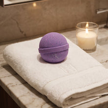A purple spherical bath bomb sits on a folded white towel on a marble bathroom counter, with a lit candle in a glass holder nearby.
