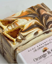 A close-up of a rectangular bar of artisan soap with a marbled swirl pattern of cream, brown, and orange, topped with dried orange peel flakes. A white paper label at the bottom reads 'RCDC ESSENTIALS An Artisan Soap Co.