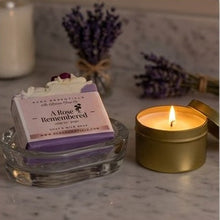 A bar of soap labeled 'A Rose Remembered' by Mood Essentials sits in a clear glass dish next to a lit gold tin candle on a marble surface, with dried lavender sprigs in the background.