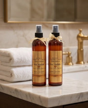 Two amber spray bottles of Pome Essentials Stay Awkward body wash with gold labels and beige ribbon bows, standing on a marble bathroom counter next to folded white towels and a gold faucet.