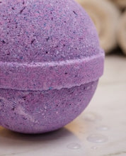 A close-up of a spherical lavender bath bomb with a textured, granular surface and a horizontal seam, resting on a light marble-like surface with water droplets.