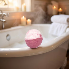 A pink and white bath bomb sits on the edge of a white bathtub with steam rising, candles lit in the background, and a folded white towel on the tub.