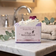 A boxed soap bar sits on a bathroom counter with a faucet, towel, and greenery in the background. The box reads 'A Rose Remembered' and shows the brand 'BCCD Essentials'.