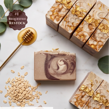 Artisan soap bars with gold leaf and honeycomb patterns, displayed on a white marble surface with eucalyptus leaves, oats, a gold honey dipper, and a '#1 REQUESTED RESTOCK' badge.