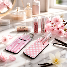 Pink lip balm cases with key rings and a lip balm tube, styled on a marble surface with pink flowers, vanilla beans, and cosmetic items in the background.