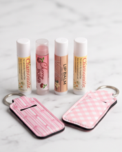 Four lip balm tubes and two pink keychain pouches arranged on a light marble surface. The lip balms are labeled Chamomile, Cherry Bomb, and Wild Berry, with two matching pink checkered and pink striped keychain cases.