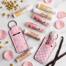 A styled flat-lay of lip balm tubes and two pink patterned key cases on a white marble surface, surrounded by pink petals, dried chamomile flowers, and vanilla beans. Visible lip balm labels include Chamomile, Butter Jelly Cherry Bomb, and Cherry Bomb.