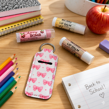 A pink bow patterned lip balm keychain holder sits on a wooden desk surrounded by school supplies, including colored pencils, notebooks, an apple, and lip balm tubes.