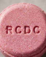 A close-up of a round pink solid shampoo or soap bar with the letters 'RCDC' embossed on the front, set against a neutral gray background.