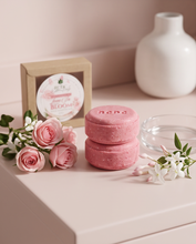 Two pink round solid shampoo  bars stacked on top of each other, displayed on a light pink surface with pink roses, a small glass dish, and a kraft box behind them. A white ceramic vase is blurred in the background.