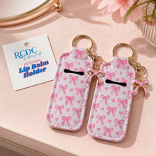 Two pink and white keychain lip balm holders with bow and heart patterns, gold keyrings, and small bow charms, displayed on a light pink surface next to a RCDC essential keychain card.
