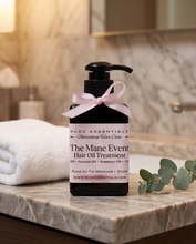 A bottle of RCDC Essentials 'The Mane Event' Hair Oil Treatment sits on a marble bathroom counter next to a folded white towel and eucalyptus leaves. The bottle is black with a black pump, featuring a white label with pink text and a light pink ribbon tied in a bow around the neck.