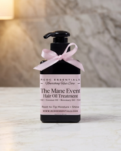 A black pump bottle of RCDC Essentials 'The Mane Event' Hair Oil Treatment, tied with a light pink satin ribbon, set on a white marble surface with a blurred marble wall background.