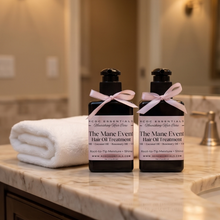Two black bottles of KCDCE Essentials 'The Mane Event' Hair Oil Treatment tied with pink ribbons, displayed on a marble bathroom counter next to a folded white towel.