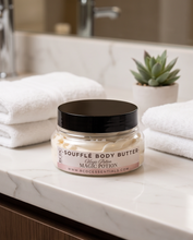A jar of RCDC Sofflé Body Butter labeled 'Magic Potion' sits on a white marble bathroom counter, flanked by folded white towels and a small potted succulent.