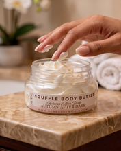 A hand scoops creamy white body butter from a clear jar labeled 'SOUFFLÉ BODY BUTTER Autumn After Dark' on a marble countertop with rolled towels and a blurred indoor setting in the background.