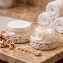 Two jars of SOUFFLÉ BODY BUTTER labeled Autumn After Dark, one closed with a white lid and one open showing the white cream, styled on a marble surface with a gold spoon and dried botanicals.