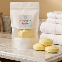A white stand-up pouch labeled 'BC15 Aromantic Shower Steamers Confidence' sits on a marble bathroom counter next to a stack of white towels, with three pale yellow shower steamers placed in front.