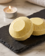 Two round pale yellow bath or body products wrapped in clear plastic, displayed on a dark slate board with a small lit candle in the background on a marble surface.