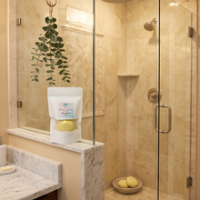 A bathroom scene showing a walk-in shower with beige stone walls and a glass door, alongside a countertop with a white bag of bath bombs and a hanging green plant.