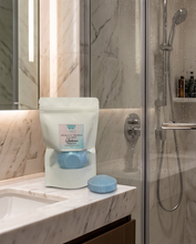 A white resealable pouch labeled 'Rose Gold' and 'Nude' sits on a marble bathroom counter next to a matching round blue bath bomb, with a glass shower door and tiled wall in the background.