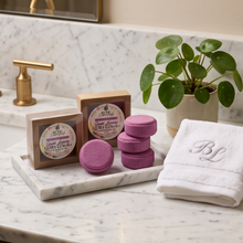 A styled bathroom countertop scene showing a boxed bath product labeled 'Royal Bath Deep Luxury Soak' alongside three round purple bath soaks and a folded white embroidered towel. A marble tray, brass faucet, potted plant, and marble wall are visible.