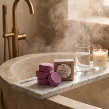 Purple solid shampoo bar in a wooden box, stacked round bath bombs on a marble tray, a glass of water, and a lit candle on a bathroom bathtub caddy.
