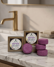 Three purple rectangular bath soaps in a matte finish, with two in their gold and white packaging and one standing loose in front, displayed on a marble bathroom counter next to a gold faucet.