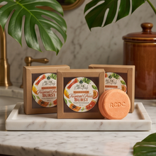 Three square kraft boxes of RCDC ESSENTIALS  Fruit Burst bath products are arranged on a white marble tray, with one round peach-colored bar soap placed in front. Large green monstera leaves and a brown ceramic jar appear in the softly lit bathroom setting.