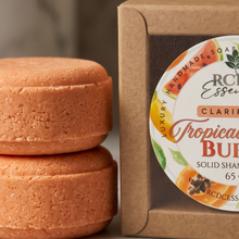 Two round orange-brown solid shampoo bars stacked on a marble surface beside a kraft paper box labeled 'R.C.