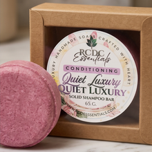 A pink solid shampoo bar in front of its kraft paper window box packaging, with the round label reading RCDC Essentials Quiet Luxury Solid Shampoo Bar.