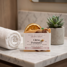 A boxed bar of soap labeled Citrus Pomander by RoCo Essentials sits on a marble bathroom counter, styled with a rolled white towel, a dried orange slice, and a small potted succulent.