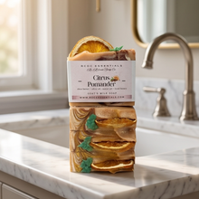 A stack of three handcrafted goat milk soaps by Rose Essentials The Gilded Spoon Co. on a white marble bathroom counter, featuring citrus pomander scent with dried orange slices and green leaves, with a blurred bathroom sink and faucet in the background.