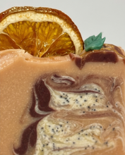 Close-up of a swirled layered dessert or soap in warm beige, brown, and cream tones with dark chocolate-like streaks and small black poppy seeds, topped with a dried orange slice and a small green leaf on a light gray background.